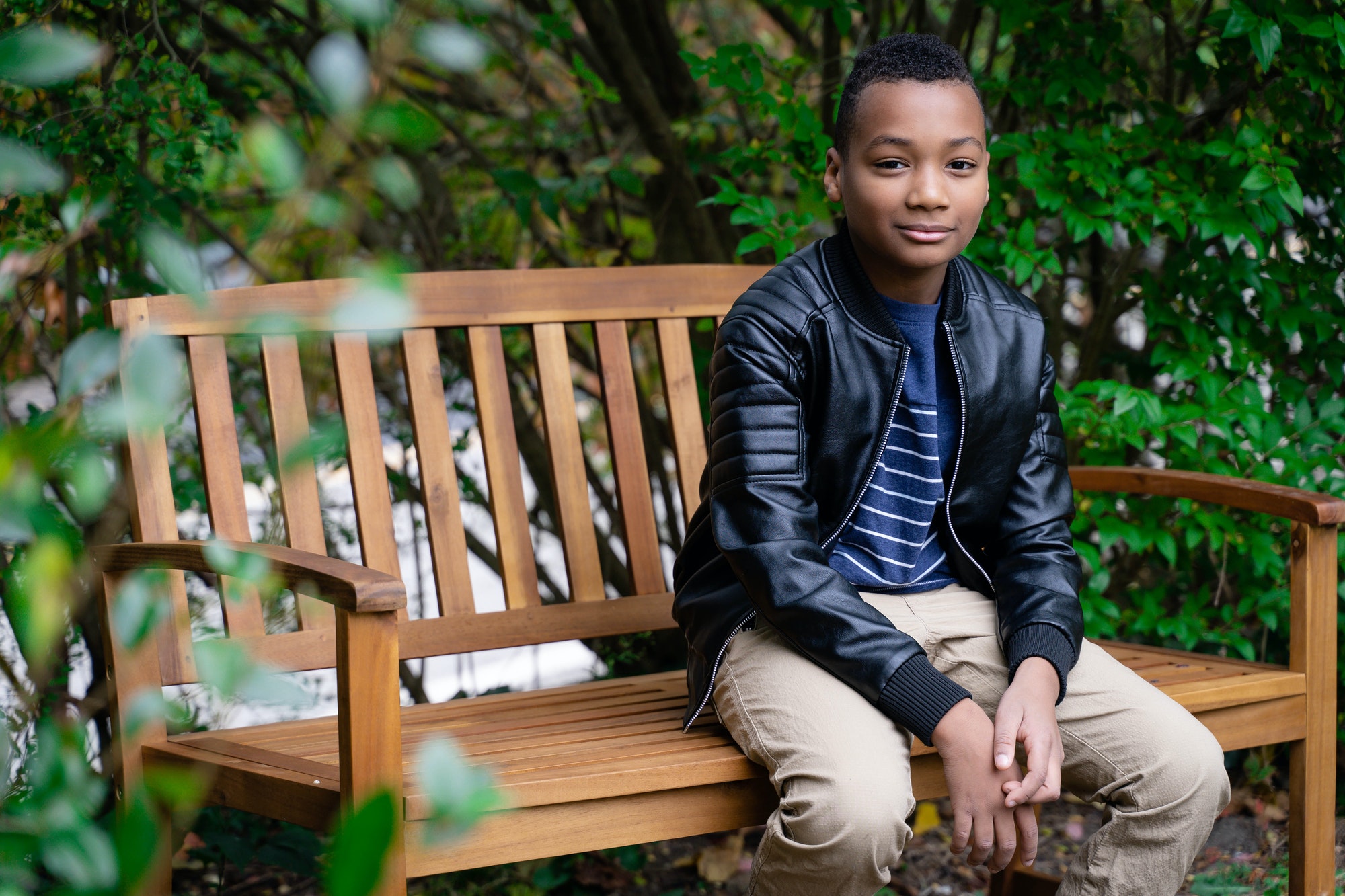 Young black boy smiling, sitting on a park bench wearing a leather jacket